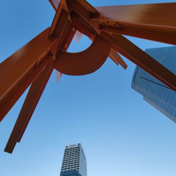 The Calling - The photograph captures a low-angle perspective of multiple modern skyscrapers, including a prominent US Bank building, set against a clear blue sky. In the foreground, there is a large orange abstract sculpture with geometric shapes and structural elements. The composition emphasizes the height of the buildings and the sculpture's bold color contrast with the sky. - Local Flavor Near Me - Milwaukee, Wisconsin