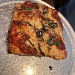 Grandma's - A slice of pizza featuring melted cheese, tomato sauce, fresh basil leaves, and grated cheese on top, placed on a round metal plate. The pizza has a slightly charred crust and is served in a dining environment, with a small container of sauce visible in the background.