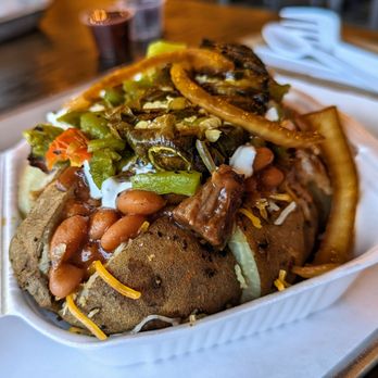 Duke City BBQ - 505 Baked Potato with pulled pork - pretty remarkably good - A baked potato is topped with chili, shredded cheese, beans, and sautéed vegetables including onions and peppers. The dish is served in a white disposable container, with a blurred background showing drinks and napkins. The toppings are abundant, covering the potato entirely, and the presentation emphasizes a hearty, flavorful meal. - Barbeque Near Me - Albuquerque, New Mexico