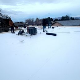 Final product - Multiple workers are on the flat roof of a residential building, installing or repairing roofing materials. The roof is white, and various tools and equipment, including rolls of roofing membrane and ventilation units, are present. The background shows neighboring houses, trees, and a cloudy sky, indicating an outdoor construction or maintenance activity.