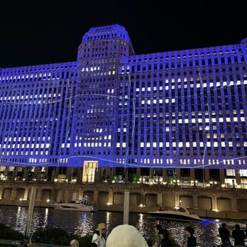 Art on theMART - The image shows the Art on theMART building in Chicago lit up with blue projection lighting at night, with people and boats along the river. - Art Installation Near Me - Chicago, Illinois