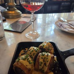 Pigs in a Blanket - A glass of red wine and a small cast iron skillet with sushi rolls topped with sauce and chopped chives on a marble table at a restaurant.