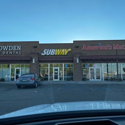 Front area - A shopping plaza with storefront signs for Subway, America’s Mattress, and Cowden Family Dental, with parked cars in front and a clear blue sky.