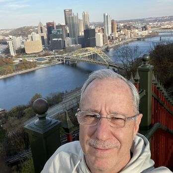 Station Square - A man with gray hair and glasses is taking a selfie on a hill or viewpoint overlooking a city skyline and river. The background features tall buildings, a bridge, and a river, with a clear sky above. The man is smiling slightly and appears to be enjoying the view. - Shopping Centers Near Me - Pittsburgh, Pennsylvania