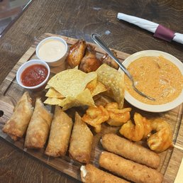 Sampler - A wooden serving tray contains a variety of fried appetizers including spring rolls, chicken wings, and shrimp coated in sauce. There are also tortilla chips and two small cups of dipping sauces, one white and one red. A bowl of orange-colored soup with a spoon is placed on the tray, and a napkin is positioned on the table nearby. The setting appears to be a restaurant with a dark wooden table.