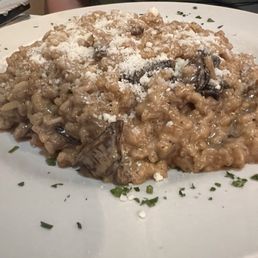 Risotto Funghi Porcini Delicious! - A serving of mushroom risotto on a white plate, topped with grated cheese and chopped herbs, served in an Italian restaurant setting.