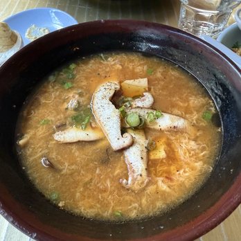 Myers & Chang - Esti's Hot + Sour Soup - A bowl of hot + sour soup with sliced pork, green onions, and egg ribbons, served in a restaurant setting with water glasses and other dishes. - Asian Fusion Near Me - Boston, Massachusetts