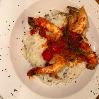 La Strada Restaurant - Risotto ai gamberi - A white plate holds risotto ai gamberi, a traditional Italian rice dish topped with cooked shrimp, tomato sauce, and garnished with fresh herbs. The risotto appears creamy with visible rice grains, and the shrimp are arranged on top, partially covered by the sauce. The plate is decorated with small green herbs around the edge, and the dish is presented on a wooden table. - Pizza Near Me - Millburn, New Jersey