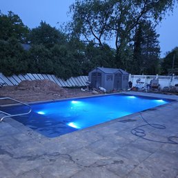 The image depicts a backyard with a rectangular in-ground swimming pool illuminated by blue LED lights at dusk. The pool is surrounded by a stone patio, and there are construction materials and tools nearby, indicating ongoing work. In the background, there are trees, a white fence, and small storage sheds, with neighboring houses visible beyond the fence.