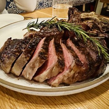 Tavernetta - O-M-G - apex option on this tight & tasty menu: 50oz bistecca alla fiorentina - A large, cooked steak is sliced and presented on a white oval plate, garnished with sprigs of fresh rosemary. The steak appears to be medium-rare, with a pink interior and a charred exterior. The presentation emphasizes the cut and seasoning, with a rustic dining setting visible in the background, including a wooden table and some dining accessories. - Italian Near Me - Denver, Colorado