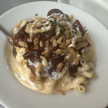 The Eagle OTR - Burnt end Mac and cheese - A serving of macaroni and cheese topped with burnt ends, crispy fried onions, and a drizzle of sauce is presented on a white plate. The dish is garnished with chopped green onions and appears creamy with a variety of textures. The setting includes a gray table surface and a white plate in the background, indicating a casual dining environment at The Eagle. - American Near Me - Cincinnati, Ohio