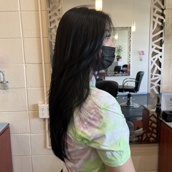 Glamor Cuts - Layered haircut - A woman with long black hair, wearing a black face mask and a tie-dye shirt, stands in front of a mirror inside a hair salon. - Hair Salons Near Me - Concord, California