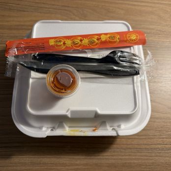Bamboo by Kulantro - 11.25.2025 - A foam takeout food container is positioned on a wooden table, with a sealed pair of chopsticks in red packaging, a small plastic container of soy sauce, and a black utensil, possibly a fork or spoon, wrapped in plastic, arranged on top of the container. - Vietnamese Near Me - Albuquerque, New Mexico
