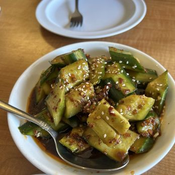 Wild Ginger - S1. Spicy cucumber salad / 拍黄瓜 - A bowl of cucumber salad with a spicy sauce and sesame seeds, served on a wooden table in a restaurant setting. - Chinese Near Me - Alameda, California