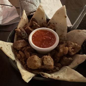 El Bruno's - A plate contains fried chicken pieces, triangular tortillas arranged around a small bowl of red salsa in the center, and a lime wedge on the side. The dish is served on a dark-colored plate, placed on a table with a napkin and some paper in the background. The setting appears to be a restaurant with wooden chairs visible in the background. - Mexican Near Me - Albuquerque, New Mexico