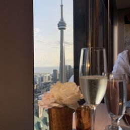 The image shows a table with a copper-colored flower pot holding a white flower, a glass of champagne, and a glass of water. The table is positioned next to a large window, through which the CN Tower in Toronto is visible against a partly cloudy sky. The interior includes a person partially visible on the right side, and the scene suggests a dining or lounge setting with a city view.