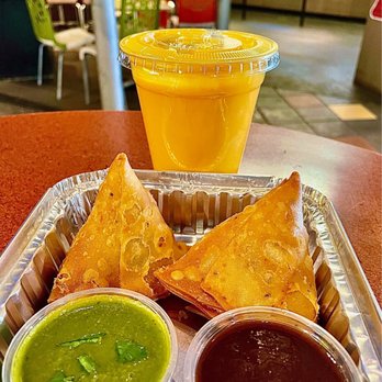 Chutney's - A tray contains two golden-brown samosas accompanied by green chutney and tamarind sauce, placed on a red table. Behind the tray, there is a plastic cup filled with mango lassi, topped with a plastic lid. The background shows a casual restaurant interior with colorful chairs and a counter area, indicating an informal dining environment. - Indian Near Me - Cambridge, Massachusetts