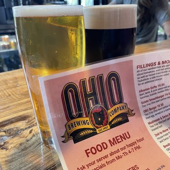 Ohio Brewing - Lil Vata & Bowsie - Two glasses of craft beer, one light-colored and one dark, are positioned on a wooden bar counter. The light beer appears golden with a foamy head, while the dark beer is nearly black with a tan foam. A menu from Ohio Brewing is partially visible in the foreground, listing food and drink options. The background shows a bar interior with shelves and glassware, indicating a brewery or pub environment. - Breweries Near Me - Columbus, Ohio