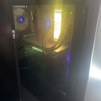 Quality Plus Computer - A black computer tower with a clear side panel reveals internal hardware components such as a graphics card labeled 'GEFORCE RTX,' RAM modules with yellow lighting, a cooling fan with blue LED, and other internal wiring. The case is positioned on a textured surface, and the internal components are illuminated with various LED lights, indicating a high-performance or gaming setup. - IT Services & Computer Repair Near Me - Chino Hills, California