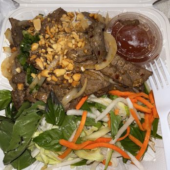 Wrap N Roll - B9. Lemongrass Beef noodle Bowl - The image shows a takeout container with a Vietnamese lemongrass beef noodle bowl. It contains sliced beef, onions, and chopped peanuts on top. The bowl also includes fresh vegetables such as lettuce, carrots, bean sprouts, and basil leaves. A small cup of sauce or dressing is placed in the container. The food appears to be a quick, flavorful Vietnamese dish emphasizing fresh ingredients. - Vietnamese Near Me - Albuquerque, New Mexico