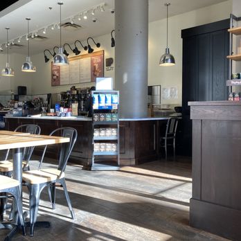 Peet's Coffee - Peet's Coffee - The image shows the interior of a coffee shop with a dark wood counter and seating area. Pendant lights hang from the ceiling, illuminating the space. A menu board is visible behind the counter, along with coffee-making equipment. Sunlight streams through windows, creating shadows on the wooden floor. The decor includes a large concrete pillar and dark wood paneling, contributing to a modern, cozy atmosphere. - Coffee & Tea Near Me - Washington, DC, District of Columbia