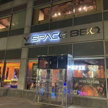 Y Space BBQ - Entrance - The image shows the exterior of Y Space BBQ, a restaurant located in a multi-story building. The restaurant's sign is illuminated, displaying the name 'Y SPACE BBQ' with a modern font. Large windows reveal the interior, which is lit with warm lighting and visible seating arrangements. The entrance features a glass door with a metallic frame, and there is a neon sign indicating the restaurant is open. The surrounding sidewalk is visible in the foreground. - Barbeque Near Me - Flushing, New York