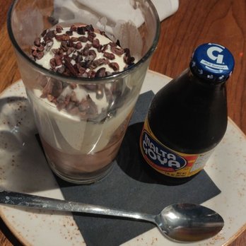 Para Maria - Malta chocolate float ($12). - A dessert drink featuring whipped cream and chocolate sprinkles in a tall glass, placed on a plate with a spoon and a bottle of Malta Hoya soda. The drink appears creamy and is garnished with chocolate sprinkles, with a spoon resting on the plate. The setting is a wooden table in a restaurant environment. - Wine Bars Near Me - Boston, Massachusetts