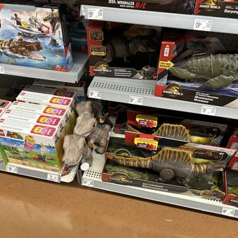 Walmart Supercenter - The way the store had the items displayed in all incorrect places yet CSM accused " you people" to me and ny children moving toys - The image shows store shelves stocked with dinosaur plush toys, including a Tyrannosaurus rex and a Spinosaurus, with some toys placed on the floor. The shelves also contain boxed dinosaur toys and other children's products. The toys are arranged in their packaging, with price tags visible. The store floor is visible at the bottom of the image, and the overall setting appears to be a toy aisle in a retail store. - Department Stores Near Me - Romeoville, Illinois