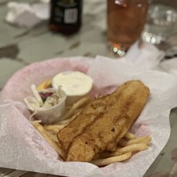Fish and Chips - A serving of fish and chips with French fries, coleslaw, and tartar sauce on a paper-lined basket, with drinks and a bottle in the background.