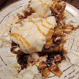 French Toast - Two slices of French toast with apple cinnamon filling are topped with whipped cream, caramel sauce, and syrup. The dish is served on a decorative white plate with a splattered design, and additional whipped cream dollops are placed around the plate. The background shows a dark countertop with a bowl and a metal container.
