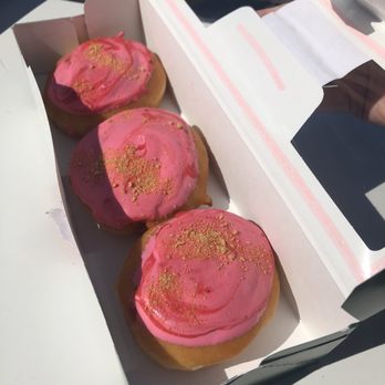 Krispy Kreme - Three donuts with pink icing and gold sprinkles are arranged in a white bakery box with a clear plastic window. The box is being held by a person with a visible hand, and the donuts are placed on a flat surface. The image is taken outdoors with sunlight casting shadows. - Donuts Near Me - Manassas, Virginia
