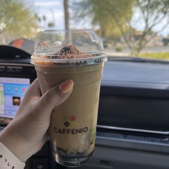 Caffenio - Cookies and cream Kfreeze - A person holds a Caffenio iced coffee with whipped topping and cocoa powder in a clear cup inside a vehicle, with a blurred outdoor background. - Cafes Near Me - Mesa, Arizona