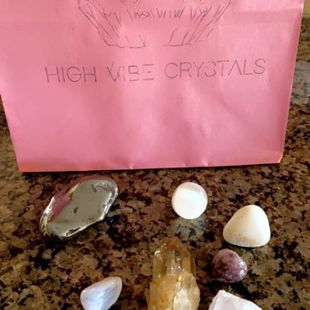 High Vibe Crystals - The image shows seven crystals of various sizes, shapes, and colors placed on a speckled granite surface. The crystals include a metallic-looking stone, a white rounded stone, a yellowish crystal, a pinkish stone, and others with different textures. In the background, there is a pink paper bag with a line drawing of crystals and the text 'High Vibe Crystals,' indicating the business related to metaphysical supplies and healing stones. - Spiritual Shop Near Me - Albuquerque, New Mexico