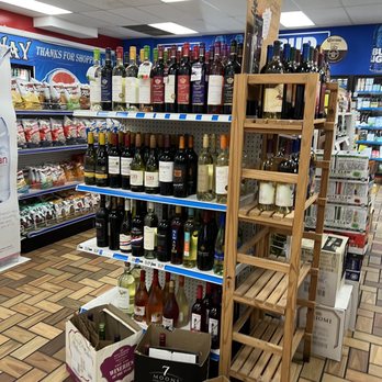 RaceWay - Wine bottles are organized on metal shelves and a wooden rack in a grocery store aisle. The shelves contain various types of wine, with some bottles on the floor in shopping bags. The store has a tiled floor, overhead fluorescent lighting, and visible snack and beverage sections in the background. - Convenience Stores Near Me - Birmingham, Alabama