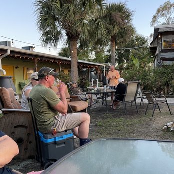 Low Key Hideaway - A group of people is seated outdoors at Low Key Hideaway in Cedar Key, Florida, with palm trees and a building in the background. Some individuals are drinking and talking, while one person stands near a table. The setting features outdoor furniture, string lights, and lush greenery, indicating a casual, relaxed atmosphere suitable for social gatherings and enjoying the natural surroundings. - Bars Near Me - Cedar Key, Florida