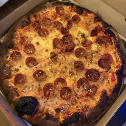 A pizza with pepperoni slices, melted cheese, and a slightly charred crust is shown in an open cardboard pizza box. The pizza appears to be freshly baked, with some burnt spots on the crust edges. The box is placed on a wooden surface, and part of a plastic bag is visible in the background.