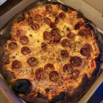 Hawt Pizza Co. - A pizza with pepperoni slices, melted cheese, and a slightly charred crust is shown in an open cardboard pizza box. The pizza appears to be freshly baked, with some burnt spots on the crust edges. The box is placed on a wooden surface, and part of a plastic bag is visible in the background. - Pizza Near Me - Albuquerque, New Mexico