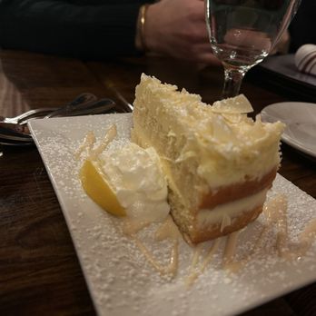 Nu Cavu - A slice of lemon cheesecake with a graham cracker crust, topped with whipped cream and a lemon wedge, is served on a white rectangular plate. The plate is dusted with powdered sugar and has a drizzle of sauce. The setting includes a wooden table, a glass of water, and utensils, with a person in a dark sweater visible in the background. - Italian Near Me - Wallkill, New York
