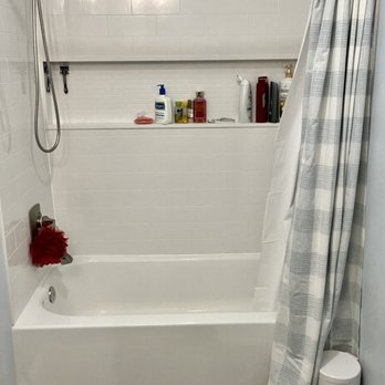 Cobalt Custom Renovations - Bath/shower combo - The image shows a bathroom with a white bathtub and a showerhead mounted on the wall. A shelf above the tub holds toiletries such as bottles of shampoo, soap, and other personal care items. A striped shower curtain partially covers the right side of the tub. The floor is covered with wood-look tiles, and a small white trash bin is placed near the corner. The walls are painted light blue, and the wall behind the tub is tiled in white. - General Contractors Near Me
