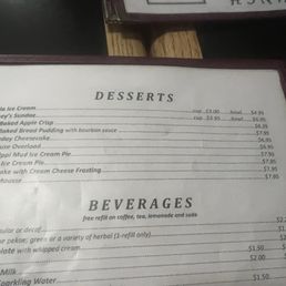 Desserts menu - The menu lists desserts including vanilla ice cream, Hershey's sundae, baked apple crisp, cheesecake, Toll House overload, Mississippi Mud Ice Cream Pie, Reese's Ice Cream Pie, carrot cake, and mocha mousse, with prices for cups and bowls. It also offers beverages such as coffee, tea, hot chocolate, milk, and sparkling water, with free refills on some drinks. The menu is printed on paper and framed, with a partial view of the surrounding environment.