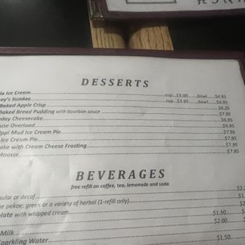 Doubleday Cafe - Desserts menu - The menu lists desserts including vanilla ice cream, Hershey's sundae, baked apple crisp, cheesecake, Toll House overload, Mississippi Mud Ice Cream Pie, Reese's Ice Cream Pie, carrot cake, and mocha mousse, with prices for cups and bowls. It also offers beverages such as coffee, tea, hot chocolate, milk, and sparkling water, with free refills on some drinks. The menu is printed on paper and framed, with a partial view of the surrounding environment. - American Near Me - Cooperstown, New York