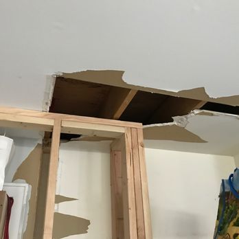 Reliable Drywall - Damage to ceiling from removing closet header beams. - The image shows a ceiling with a significant hole, revealing wooden beams and insulation inside the ceiling cavity. The surrounding ceiling surface appears damaged, with torn drywall edges. Construction materials, including a wooden frame and a bag with a colorful pattern, are visible in the room, indicating ongoing repair work. - Drywall Installation & Repair Near Me - Willow Grove, Pennsylvania