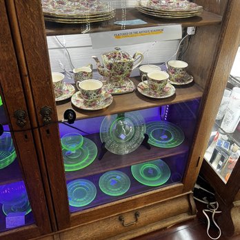 Blackberry Patch Antiques - Uranium glassware inside a display case. - A wooden display cabinet features vintage uranium glassware on the lower shelves, illuminated with a purple light that makes the glassware glow green. The upper shelves hold floral-patterned teacups, saucers, and teapots, arranged neatly. A sign indicates the items were featured on the antique roadshow. The cabinet is plugged into an electrical outlet, and the surrounding area includes other antique items and furniture. - Antiques Near Me - Powell, Ohio