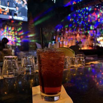 Atomic Billiards - A tall glass containing a dark-colored cocktail with ice, garnished with a lime wedge, is placed on a napkin on a bar counter. The background features upside-down glasses, colorful light reflections, and a bar area with a TV screen showing a sports event. The setting appears to be a dimly lit bar or nightclub with vibrant lighting effects. - Pool Halls Near Me - Washington, DC, District of Columbia