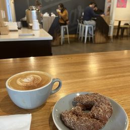 A wooden table in a coffee shop holds a latte in a white ceramic cup and a cinnamon sugar donut on a small plate. In the background, customers are seated at a counter and tables, with a dark wall and artwork visible. The setting appears cozy and modern, with natural light coming from the left.