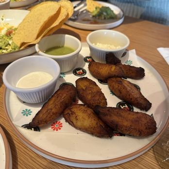 Meximodo - plantains - Fried plantains are arranged on a white plate with three small bowls of sauces, in a restaurant setting with other dishes visible. - Mexican Near Me - Metuchen, New Jersey