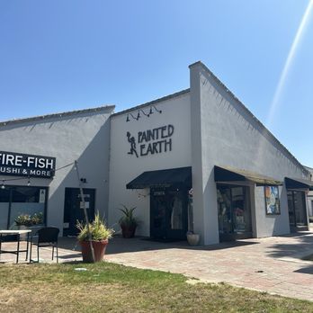 Painted Earth - Painted Earth - The image shows the exterior of Painted Earth, an art studio in Temecula, California. The building has a white facade with the Painted Earth logo and sign prominently displayed. Large windows with black awnings are visible, along with potted plants near the entrance. An outdoor seating area with chairs and tables is situated on a brick-paved sidewalk, with a small grassy area in the foreground. The sky is clear and blue, indicating a sunny day. - Paint & Sip Near Me - Temecula, California