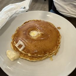 Pancakes - A white round plate holds two stacked pancakes with a pat of melting butter on top and some syrup. The pancakes are golden brown and appear fluffy. The plate is on a dark table, with part of a person's torso and arm visible in the background, and a napkin with utensils nearby.