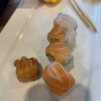 Wasabi Bistro - Rainbow Roll - The image shows a white rectangular plate with four sushi rolls topped with slices of salmon and avocado, arranged in a line. A piece of fried food, possibly tempura, is placed beside the sushi. In the background, there are chopsticks, a lemon wedge, and some shredded cabbage or similar garnish. The setting appears to be a restaurant, likely serving Japanese cuisine. - Japanese Near Me - Hershey, Pennsylvania