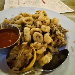 Fried Calamari - A white plate holds a serving of fried calamari garnished with chopped herbs, accompanied by a small metal cup of marinara sauce, a grilled lemon half, and a metal spoon. The plate is on a marble table with a restaurant menu, a glass of water with a straw, and a black object in the background.